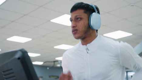 portrait view of man in white top running on treadmill indoors wearing headphones, focused expression under bright ceiling lights, gym equipment and blurred exercisers in background
