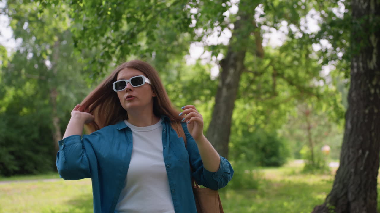 White chic adjusts hair while putting on sunglasses in green park, dressed in blue shirt and white top, showing natural confidence and casual charm surrounded by summer greenery