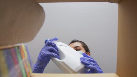 Woman-in-Mask-Taking-Cleaning-Supplies-From-Box.home-delivery,-shipping-and-pandemic-concept-woman-in-protective-medical-mask-and-gloves-opening-parcel-box,-looking-inside-and-taking-cleaning-supplies-out