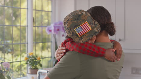 Madre-Del-Ejército-Americano-En-Uniforme-En-Casa-De-Permiso-Abrazando-A-Su-Hijo-Con-Su-Gorra-En-La-Cocina-Familiar