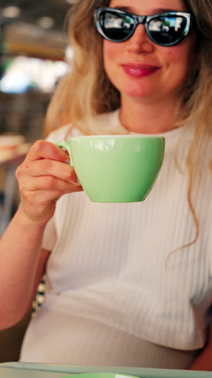 Close up of a blonde woman in a white T-shirt drinking a matcha latte out of a green cup at a terrace. Vertical