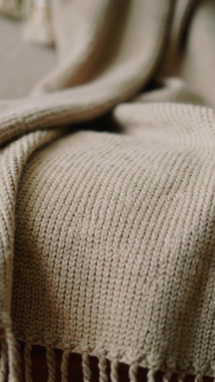 Vertikales Video: Kamera zieht den Fokus und enthüllt eine beige Kuscheldecke auf dem Sofa, die Fransen und Strickmuster zeigt.