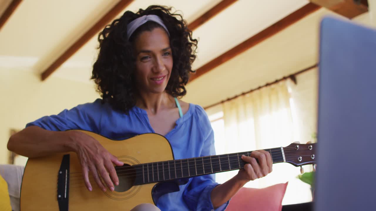 Kvinde af blandet etnicitet spiller guitar, mens hun sidder på sofaen foran sin bærbare computer derhjemme.