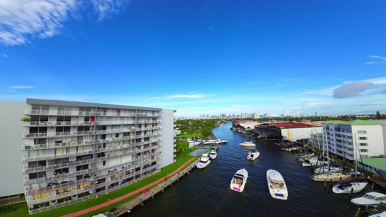 Numerous boats and yachts are standing or sailing by the river. FPV drone flies over the city scenery turning 180 degrees.