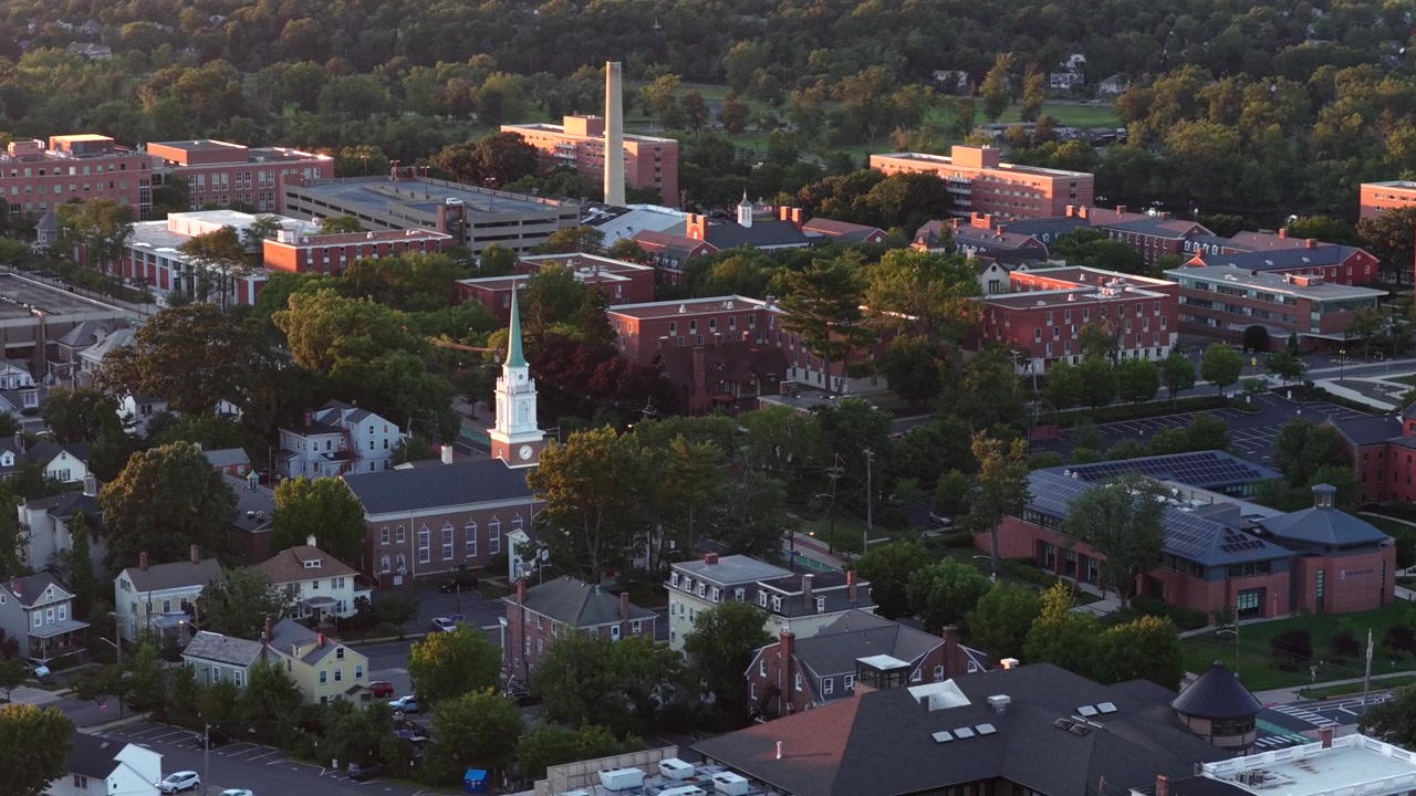 Hvitt kirketårn med klokke reiser seg blant institusjonsbygninger i rød murstein i New Brunswick, New Jersey, tett grønt trekronetak gløder varmt i gyllen morgensol. Bredt dronebilde fra luften