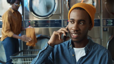 Young-Joyful-Handsome-Guy-In-Yellow-Hat-Talking-On-Mobile-Phone-And-Smiling-In-Laundry-Service-Room