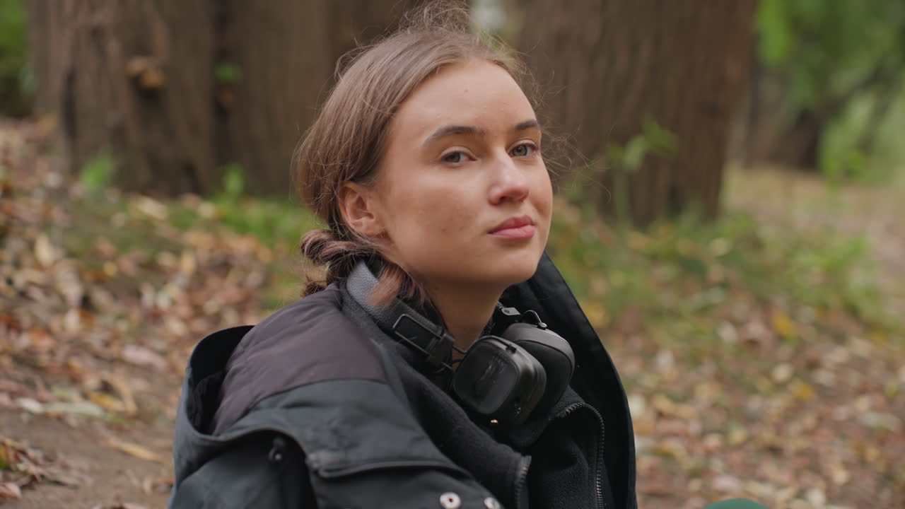 Ung kvinne ved en bekk iført hodetelefoner, ser gjennom trærne med et ettertenksomt uttrykk, lagdelt jakke og skjerf, nedfalne blader på gjørmete sti, en grunn bekk som reflekterer en trebro, myk og naturlig