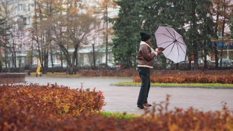 female holding umbrella, wearing knit cap, brown shearling jacket, black trousers, happily spinning umbrella in light snowfall, trees in background, enjoying autumn day, joyful and carefree in nature