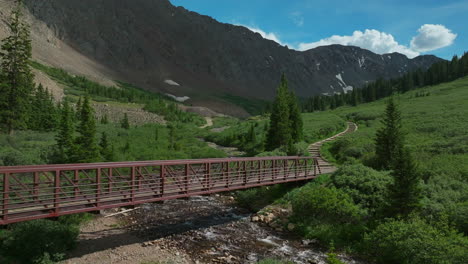 Aerial-cinematic-drone-early-morning-trailhead-bridge-over-river-Grays-and-Torreys-14er-Peaks-Rocky-Mountains-Colorado-stunning-landscape-view-mid-summer-green-beautiful-snow-on-top-backward-movement