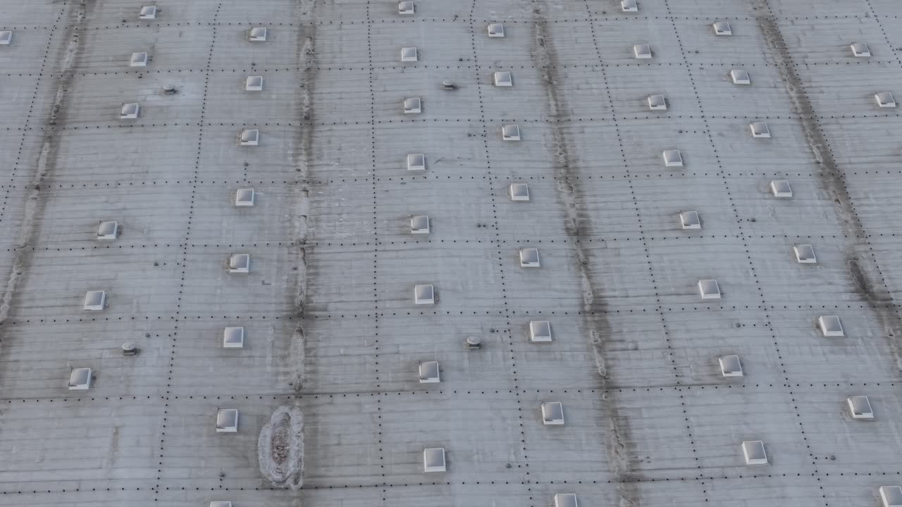 Aerial drone footage flying low over a large logistics warehouse rooftop with rows of skylights and ventilation structures. Industrial roof surface with metal framework creates a repetitive geometric