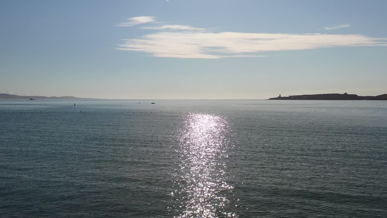 Cinematic drone shot flying over the shimmering Atlantic Ocean waters with the bright sun path reflecting on the surface and the distant coastline of Essaouira on the horizon