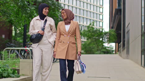 two muslim businesswomen wearing hijabs with business suits talking as they walk to work past modern office buildings