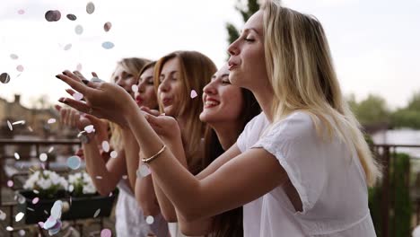 side view. attractive, smiling girls stand in a row and blow confetti from the hands. hen party concept. standing outside, terrace. view of flying bright coloured confetti. slow motion