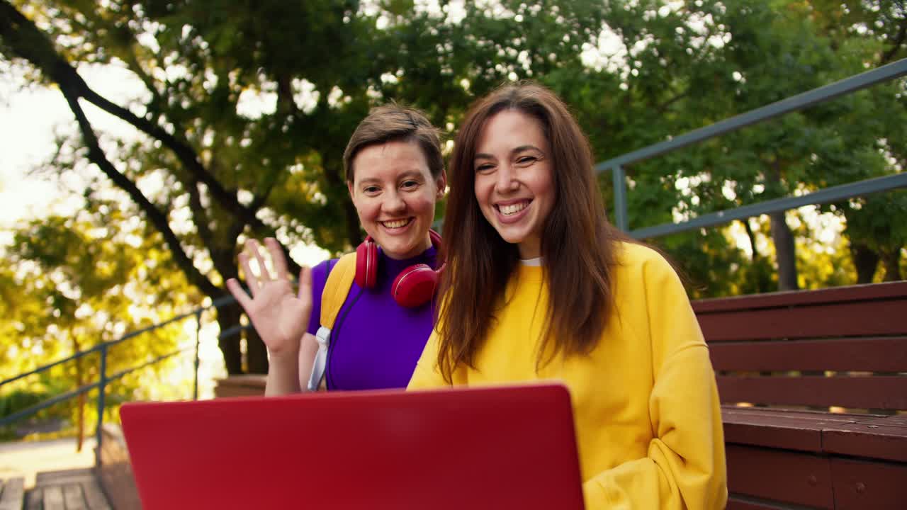 una chica morena con un suéter amarillo y una chica con un corte de pelo corto en una camiseta púrpura se sientan en puestos marrones y saludan a sus colegas en una conferencia en línea en el parque en verano