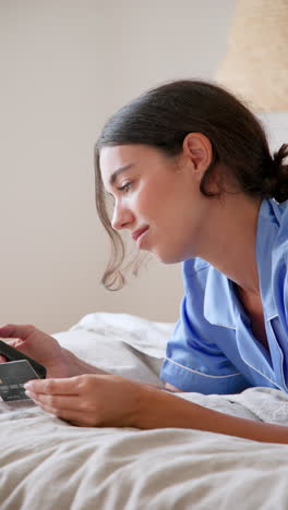vertical video: woman in pajamas smiling while online shopping with credit card in bed