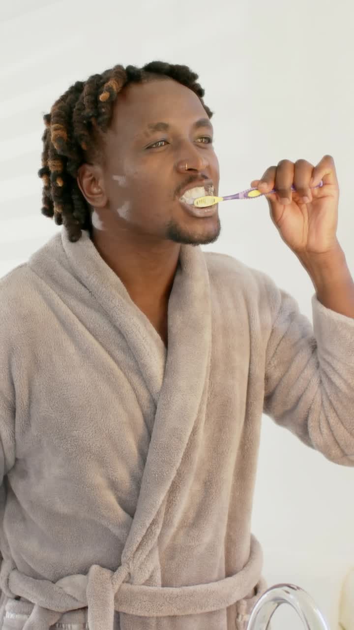 Vertical video: Raising purple toothbrush, man in robe brushing teeth at bathroom sink for grooming
