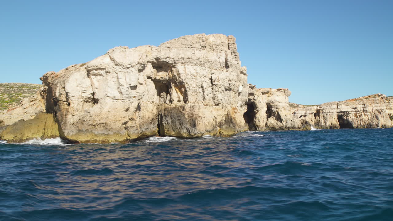 POV boat: journey on deep blue ocean sea water past rocky limestone Malta rocky rough scenic cliffs on cloudless blue sunny sky day, low angle pan