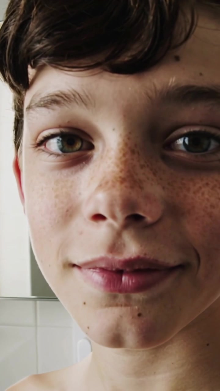 Video verticale: Adolescente a torso nudo che si gira verso la telecamera in movimento in un bagno piastrellato, mantenendo il contatto visivo.