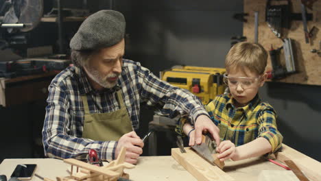 viejo carpintero caucásico con barba gris enseñando a su nieto a trabajar con madera dura en el taller