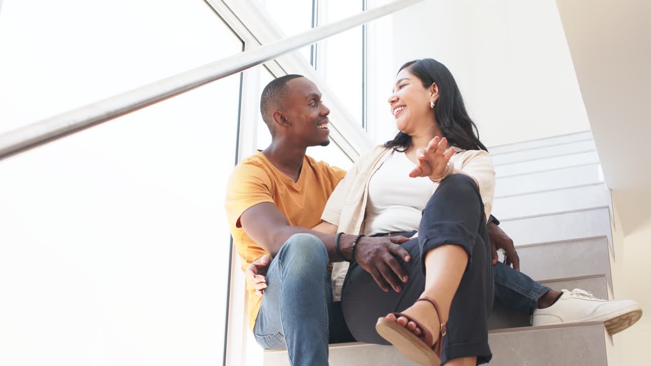 Diverse couple exchanging eye contact, bonding, laughing and leaning on stairs by large window