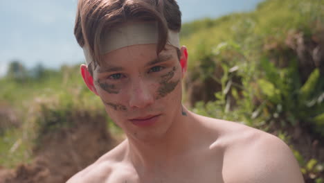 shirtless young soldier warrior in headband with muddy face stripes looking into camera with subtle smile and muzzle hand gesture, blurred riverbank and grass background under soft sunshine