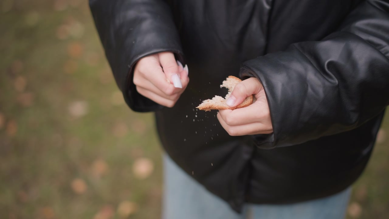 Primo piano di una donna con giacca di pelle nera che spezza il pane in mano. Le mani manipolano l'alimento mostrando i dettagli della texture su uno sfondo esterno con erba verde e foglie cadute. Morbido