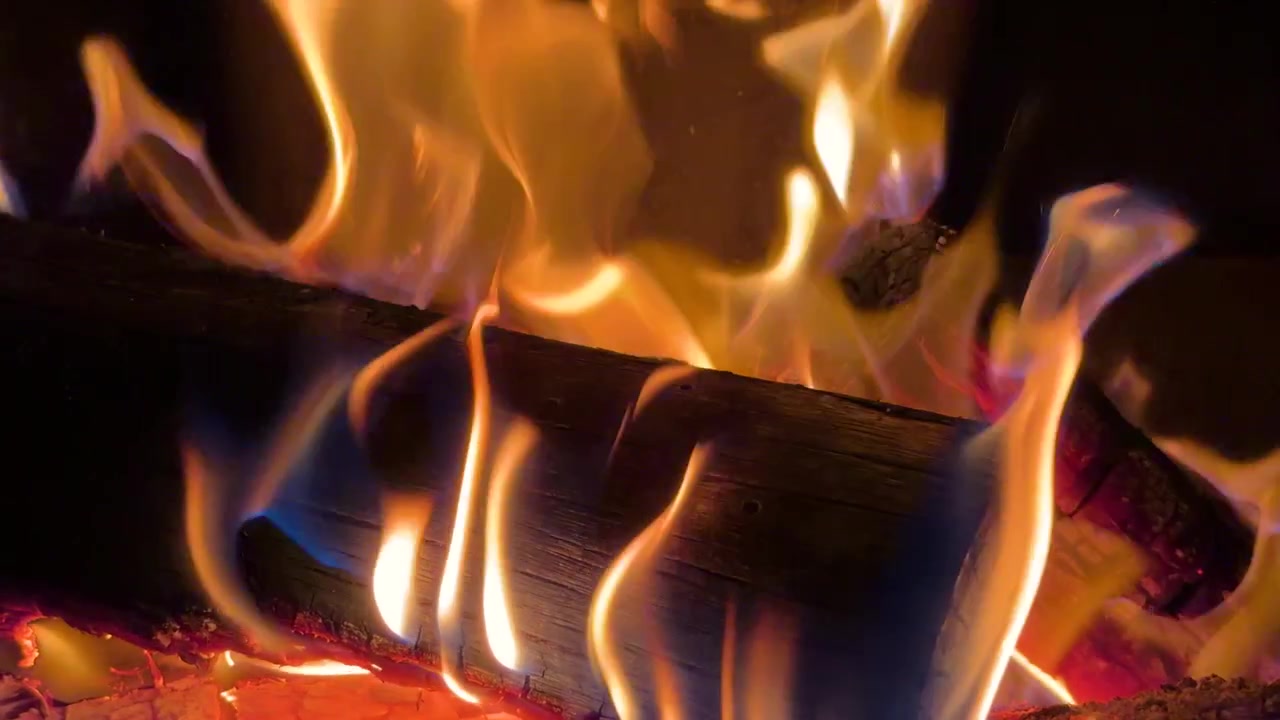 Orange flammer flimrer omkring forkullede træstammer i en hyggelig indendørs pejs med varm belysning.