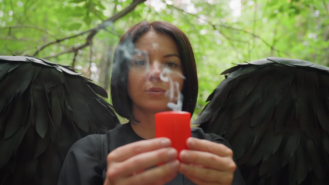 Woman dressed in black with large black wings holding burning red candle and gently blowing it out in forest, smoke rising in air symbolizing mystery, silence, and ending of dark spiritual ritual