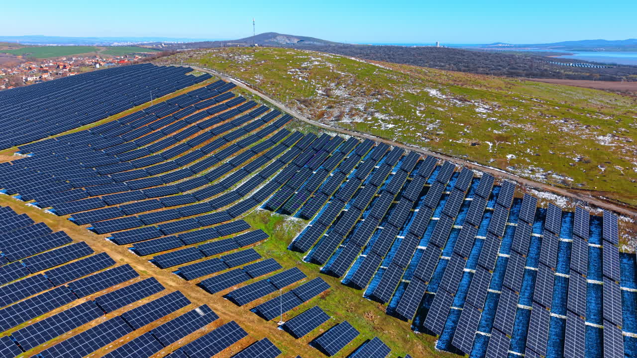 Luftfoto af stort solcelleanlæg på grønne bakker. Bred droneoptagelse af solcellepaneler installeret på græsklædte bakker under klar blå himmel med fjern havudsigt.