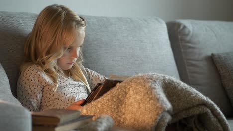 young blonde girl sitting on couch with focus to interesting book, handheld view