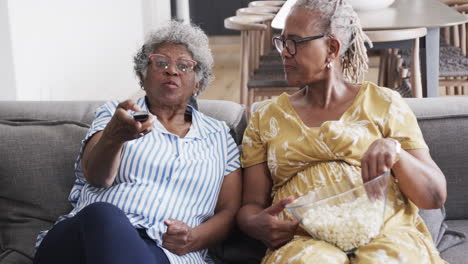 happy senior african american female friends on couch eating popcorn, using tv remote, slow motion