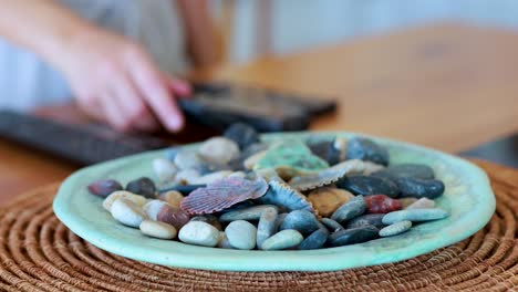 a hand reaches for and picks up a television remote control from a wooden table next to a decorative plate with stones and shells, in soft natural lighting