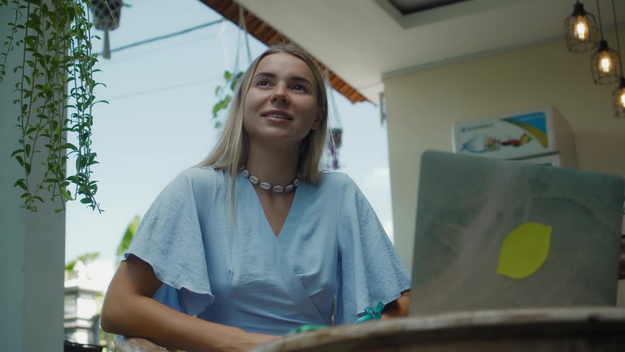 una chica en un vestido azul remotamente en línea trabajando detrás de una computadora portátil y mirando a la pantalla el patio trasero con plantas verdes