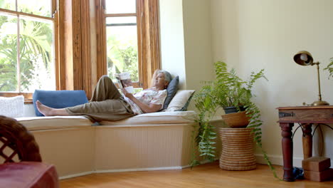 Glückliche-ältere-Frau-Mit-Gemischter-Abstammung-Auf-Der-Couch,-Die-Zu-Hause-Am-Fenster-Ein-Buch-Liest,-Zeitlupe