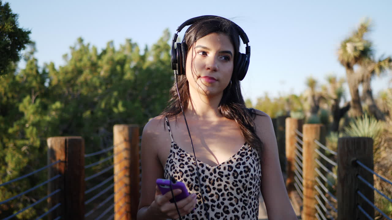 una hermosa joven hispana escuchando música pop en sus auriculares y teléfono inteligente mientras camina en cámara lenta al aire libre bajo la luz del sol