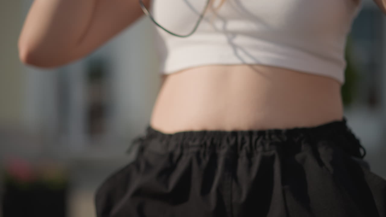 white street dancer plugging earbuds closeup, hands connecting phone to wired headphones, crop top midriff and highwaist shorts catching warm sunlight, wristbands and delicate finger gestures, urban