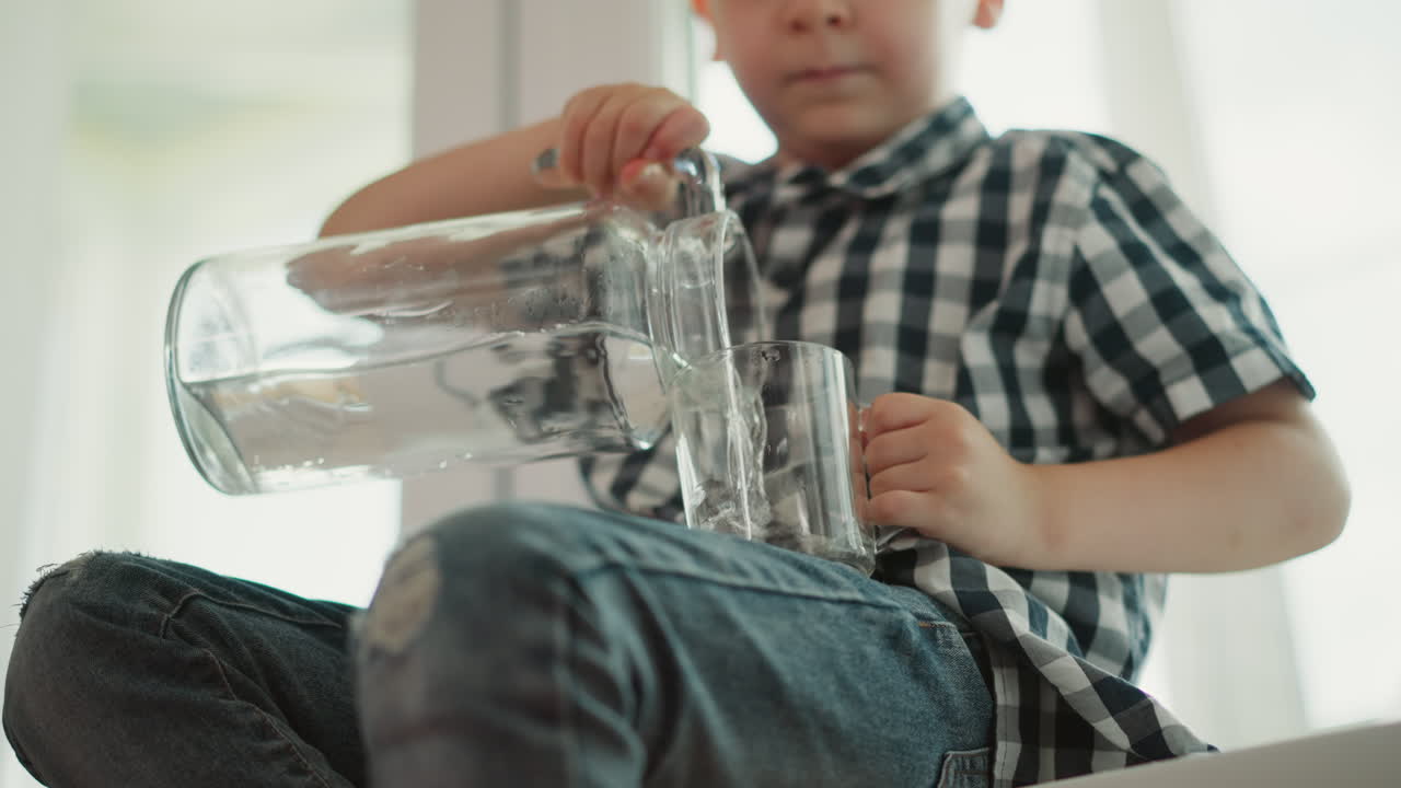 Close up upward view of kid seated on sill turning water from glass jar in right hand into cup in left hand looking at cup before lifting to drink in bright natural daylight indoors