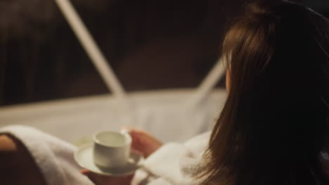 brunette woman dressed in white bathrobe relaxes relishing hot coffee in dimly lit living room. lady focuses on thoughts sitting alone in tranquil atmosphere