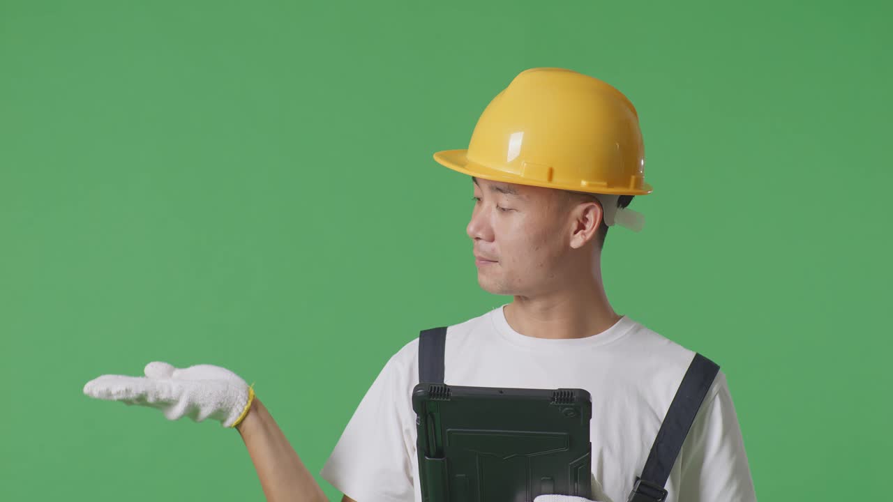 Close Up Of Asian Man Painter Wearing Safety Helmet Using A Tablet And Pointing To Side While Standing In The Green Screen Background Studio