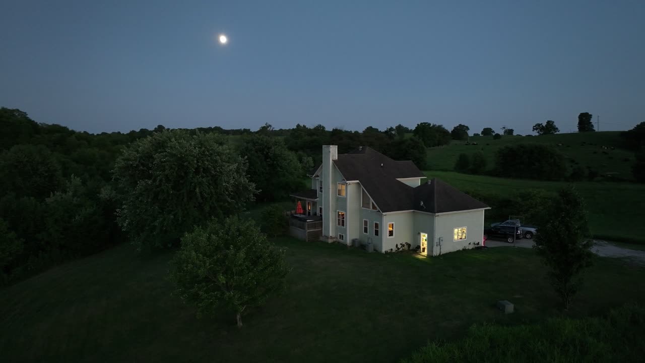 large two-story home with warm lighting on a hill in Kentucky farmland during blue hour with the moon in the background and lush greenery AERIAL ORBIT