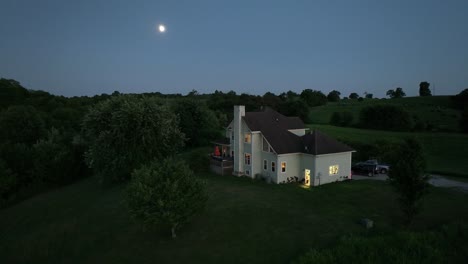 Großes-Zweistöckiges-Haus-Mit-Warmer-Beleuchtung-Auf-Einem-Hügel-In-Kentucky-Farmland-Während-Der-Blauen-Stunde-Mit-Dem-Mond-Im-Hintergrund-Und-üppiger-Grüner-Luftumlaufbahn