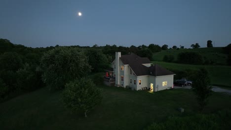 Gran-Casa-De-Dos-Pisos-Con-Iluminación-Cálida-En-Una-Colina-En-Tierras-De-Cultivo-De-Kentucky-Durante-La-Hora-Azul-Con-La-Luna-Al-Fondo-Y-Una-órbita-Aérea-De-Exuberante-Vegetación