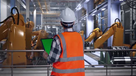 back view of asian male engineer with safety helmet working on a green screen tablet and looking around while standing with industrial robotic arms working