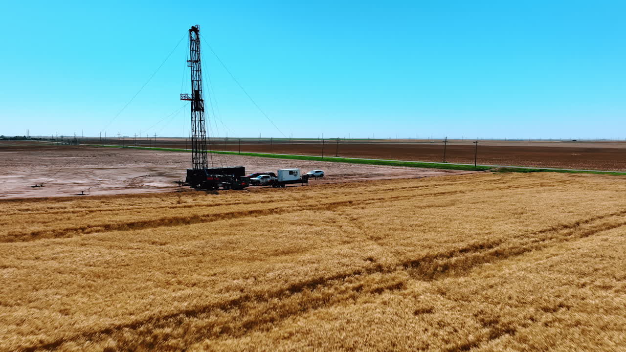 Flying over the ripe wheat field at daytime. Approaching few trucks and a drilling derrick for oil production.