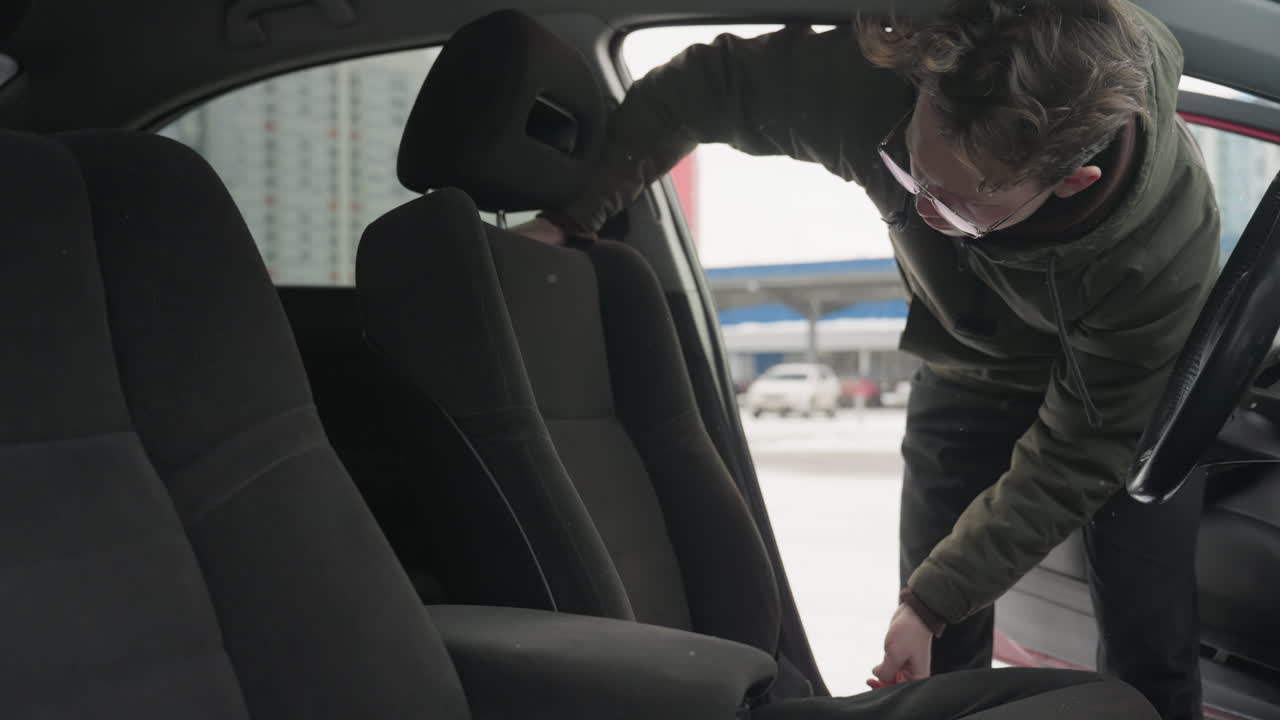 man in green winter jacket adjusts front seat of parked car by pulling it forward while standing outside in snowy weather, with cars and buildings blurred in background