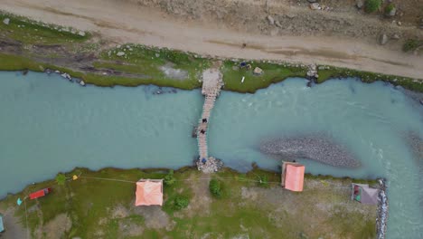 Vista-Aérea-De-Pájaro-De-Personas-Cruzando-Un-Puente-De-Madera-Sobre-El-Río-En-El-Valle-De-Basho,-Skardu