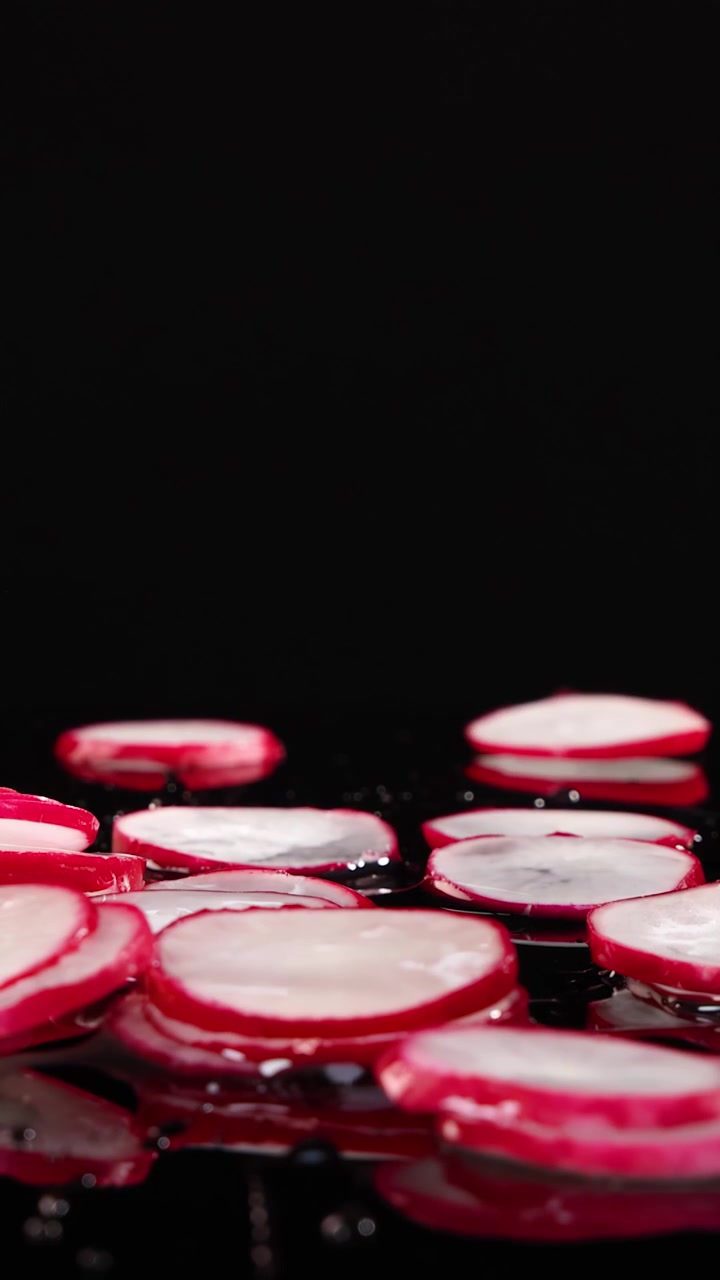 Thinly sliced radishes fall and scatter on a glossy black surface, splashing water under dramatic studio lighting with a static, close-up camera angle