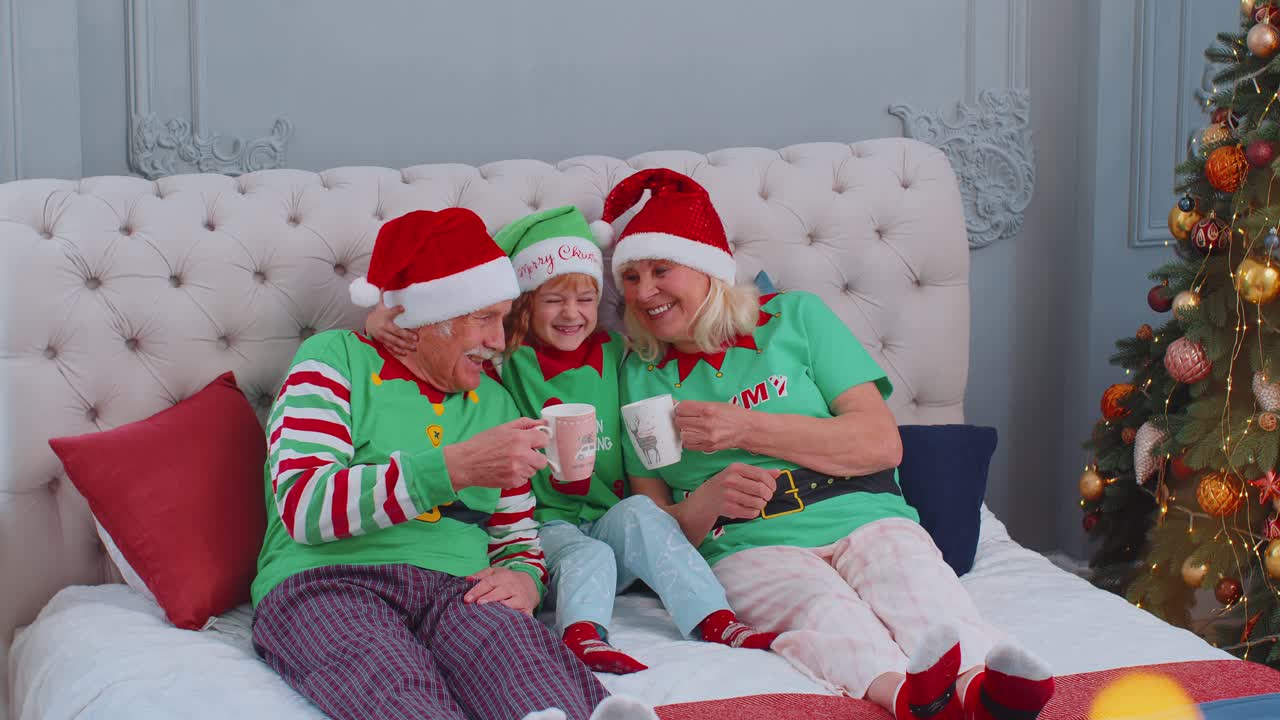 Senior couple grandparents drinking hot tea on bed with granddaughter toddler kid christmas morning