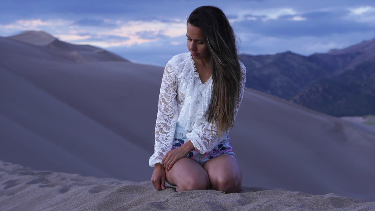 Attrice modella donna seduta in cima al Parco Nazionale Great Sand Dunes del Colorado, che riflette, guarda in basso, disegnando con il dito il paesaggio montano suggestivo al crepuscolo viola. Avventura, Montagne Rocciose, movimento statico.