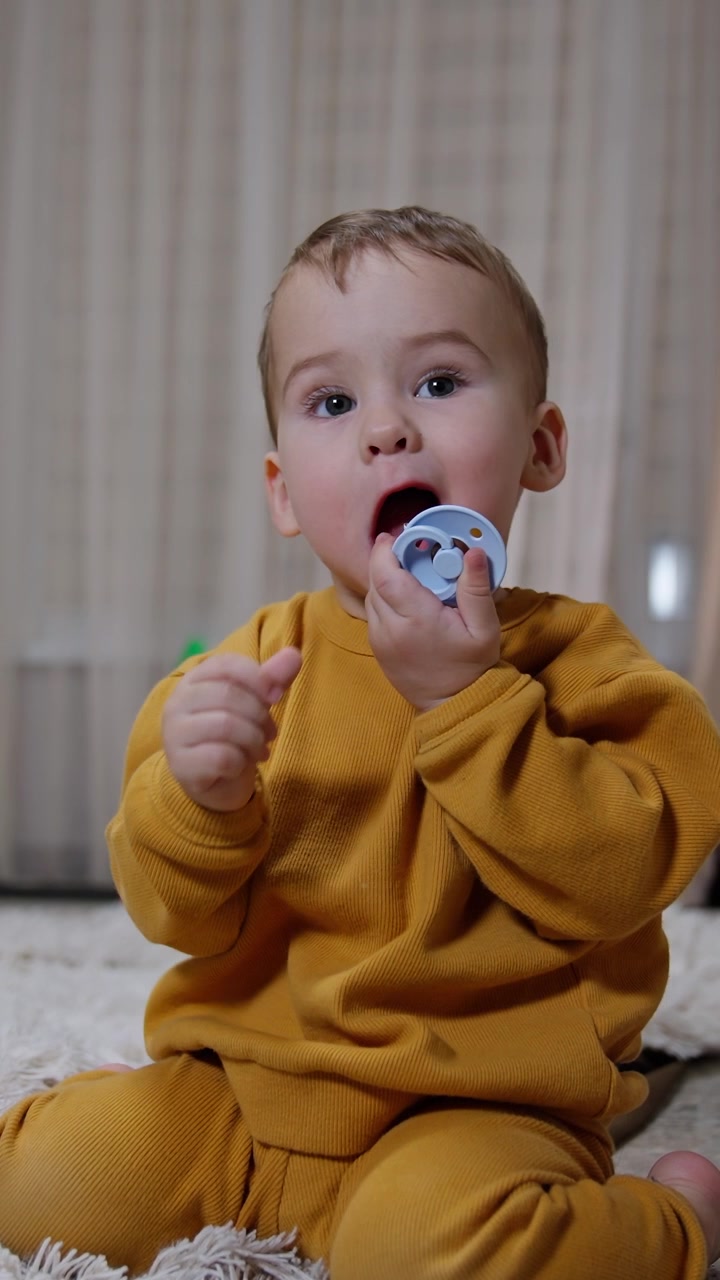 Cute baby boy sitting on the carpet holding and chewing a pacifier. Calm peaceful kid in the living room. Vertical video